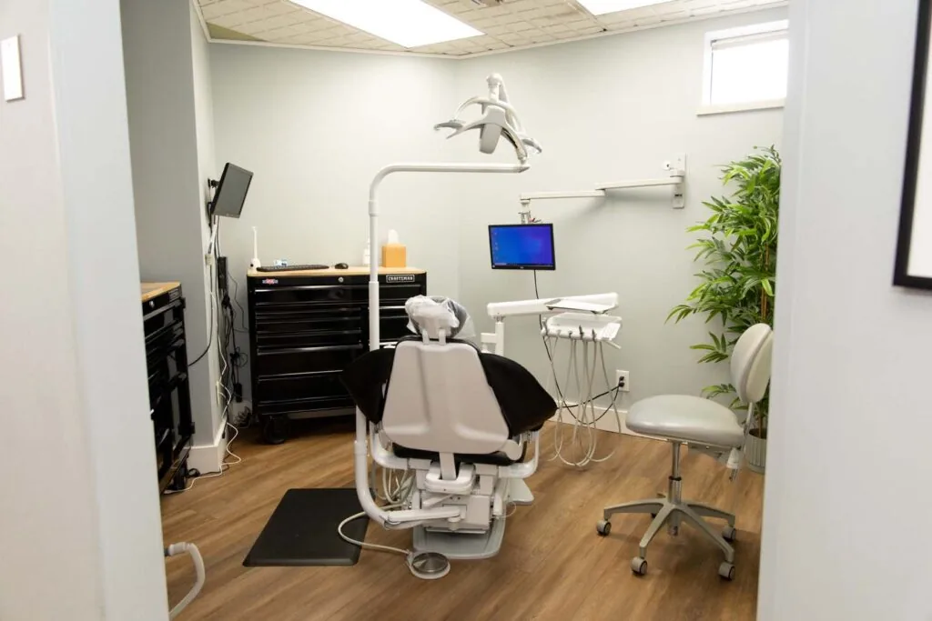 A dental office with a central dental chair equipped with overhead lights and attached instruments. The room includes a black rolling cabinet, wall-mounted computer monitor, countertop accessories, and a potted plant in a clean, professional setting.